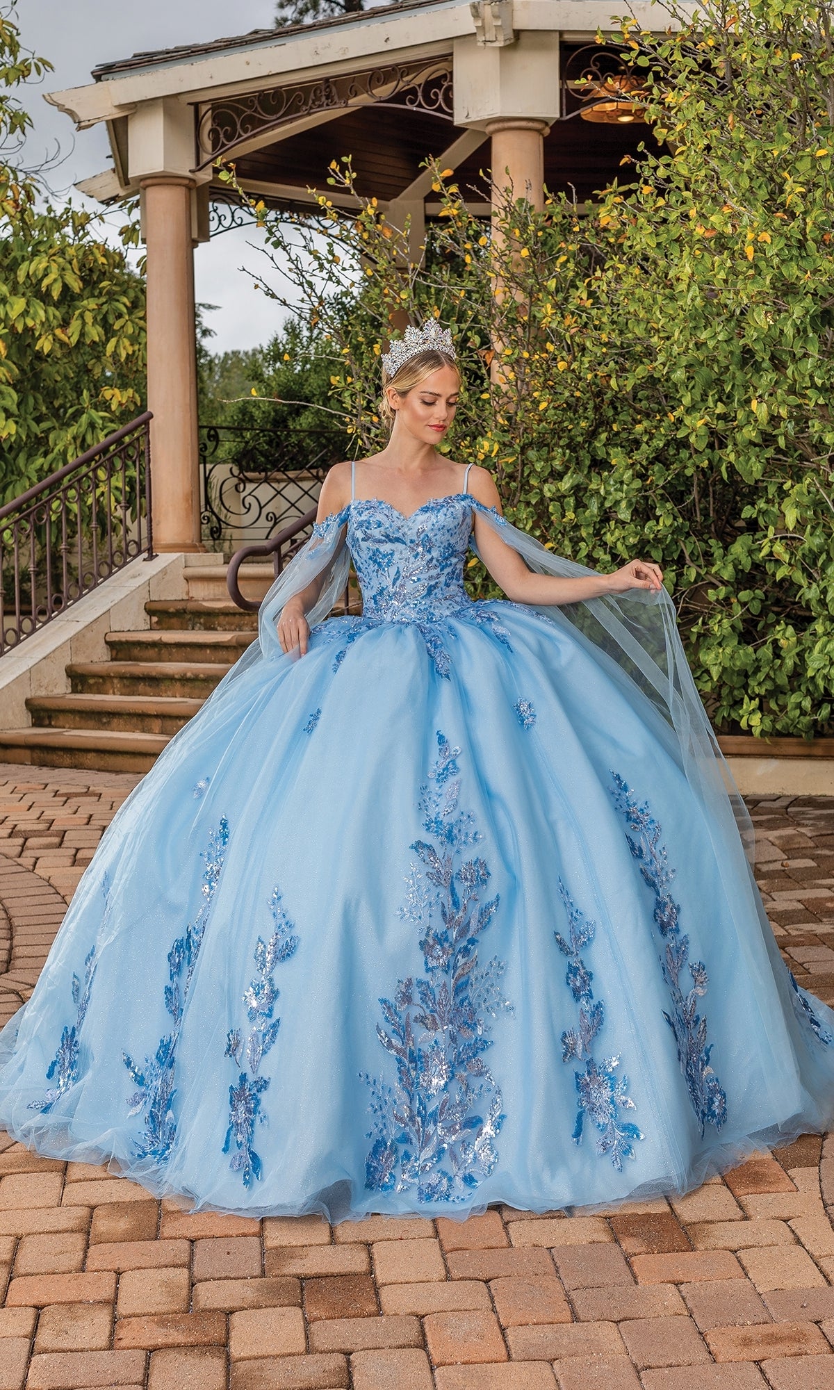 A woman stands outdoors on a brick path in front of steps and lush greenery, wearing Quinceanera Dress 1865 By Dancing Queen—a blue ball gown with floral appliqués, off-the-shoulder sleeves, and a tiara.